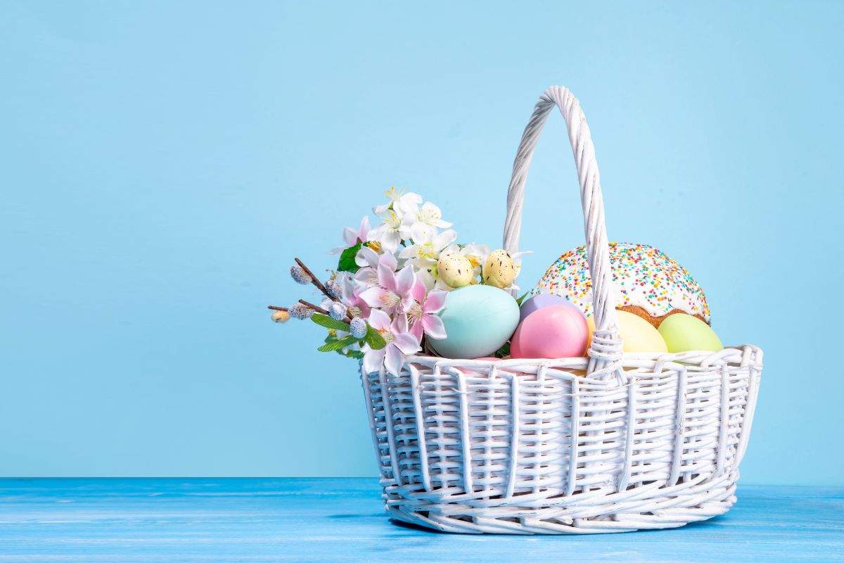 A white wicker basket filled with colourful Easter eggs, flowers, and a traditional sprinkled cake against a blue background.