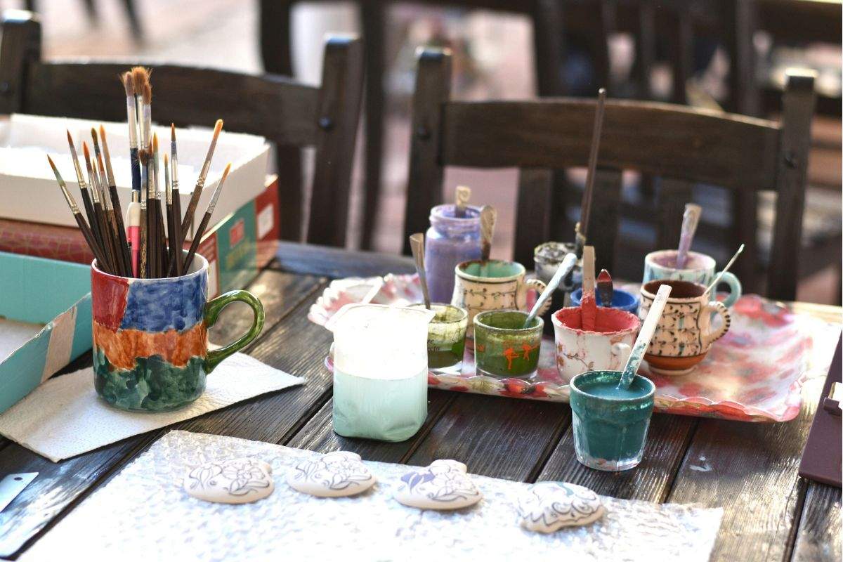 An art workstation featuring paintbrushes in a mug, small jars of colourful paint, and ceramic pieces ready to be decorated.